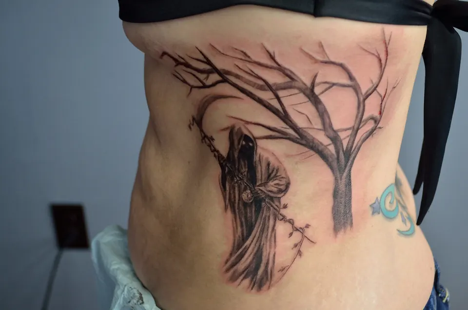 Black and grey grim reaper standing beside a dead tree on the ribs